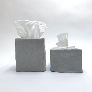 May include: Two gray linen tissue box covers. The larger cover has a white tissue box inside. The smaller cover has a white tissue box inside.