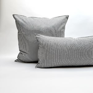 Puede incluir: Dos cojines decorativos con un patr&oacute;n de rayas blancas y negras. Un coj&iacute;n es cuadrado y el otro rectangular. Los cojines est&aacute;n hechos de una tela suave y est&aacute;n colocados sobre una superficie blanca.