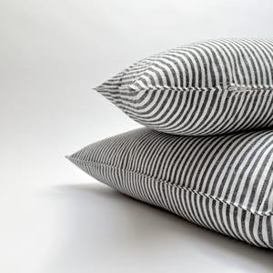Puede incluir: Dos cojines decorativos con un patr&oacute;n de rayas blancas y negras. Las almohadas est&aacute;n apiladas una encima de la otra, con una cremallera visible en la almohada superior. Las almohadas tienen un aspecto suave y texturizado.