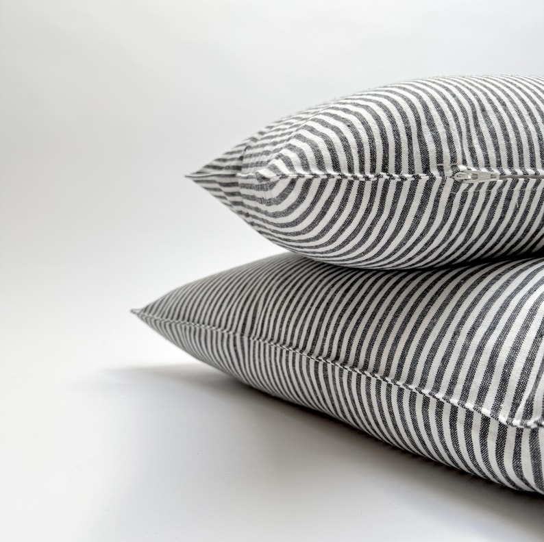 Puede incluir: Dos cojines decorativos con un patr&oacute;n de rayas blancas y negras. Las almohadas est&aacute;n apiladas una encima de la otra, con una cremallera visible en la almohada superior. Las almohadas tienen un aspecto suave y texturizado.