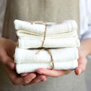 Puede incluir: Una pila de cuatro paños de cocina plegados de color blanco roto con un tejido texturizado, sujetos con un cordel marrón y una pequeña etiqueta de papel circular. Los paños están apilados cuidadosamente en forma cuadrada.
