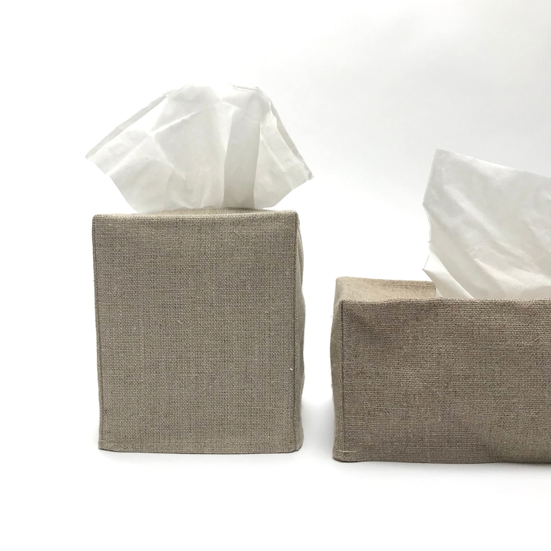 May include: Two square tissue box covers made of a natural linen fabric. The covers are in a light beige color and have a subtle woven texture.