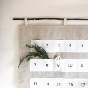 Puede incluir: Calendario de adviento de tela beige con bolsillos blancos numerados del 1 al 17. Una ramita de follaje verde y un pequeño conejito de peluche están en los bolsillos. El calendario cuelga de una vara de madera con lazos de tela beige.