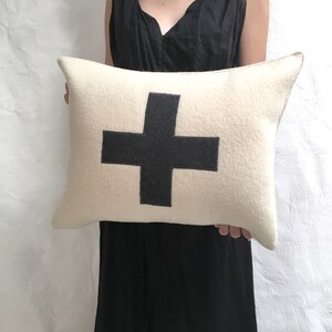 Puede incluir: Una almohada blanca con un gran signo m&aacute;s negro en el centro. La almohada est&aacute; hecha de un material suave y esponjoso.