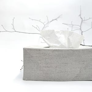 May include: A rectangular tissue box in light grey fabric, with white tissues emerging from the top. The background is white, featuring a blurred, minimalist branch design.