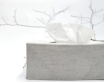 May include: A rectangular tissue box covered in light gray fabric. White tissues are pulled from the top opening. The background is white with a blurred, minimalist branch design.