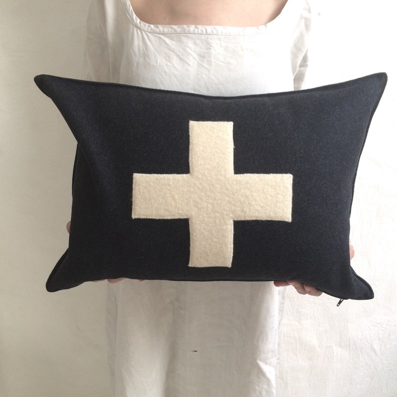 Puede incluir: Una almohada azul oscuro con una cruz blanca de fieltro en el centro. La almohada est&aacute; hecha de una tela suave y mullida.
