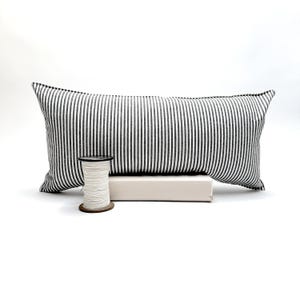 Puede incluir: Un coj&iacute;n rectangular con un dise&ntilde;o de rayas blancas y negras. Delante del coj&iacute;n hay una bobina de hilo blanco, y debajo hay un objeto peque&ntilde;o, rectangular y blanquecino.