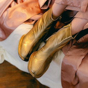 Op de afbeelding: Een paar metallic gouden enkellaarsjes met een textuur in het leer, een ronde neus en een ritssluiting aan de zijkant met een goudkleurige trekker. De laarsjes hebben een zichtbare naad over de wreef en een platte zool.