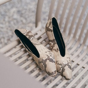 Op de afbeelding: Een paar pumps met een spitse neus en een slangenprint in beige, crème en donkerbruine tinten. De schoenen hebben een diepe V-vormige uitsnijding bij de wreef, een lage hak en een effen zwarte voering aan de binnenkant voor een strak ontwerp.