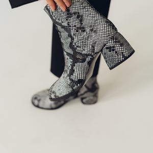 Peut inclure: Bottines à motif peau de serpent noir et blanc avec une finition texturée. Les bottines présentent un bout arrondi, un talon bloc et une hauteur mi-mollet. Le matériau affiche un motif répétitif en forme d'écailles sur toute la surface.