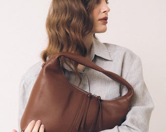 May include: A brown leather hobo bag with a pebbled texture, a single integrated shoulder strap, and a top zipper closure featuring two long leather pull tabs. The bag has a curved silhouette and is held against a light-colored long-sleeve shirt.