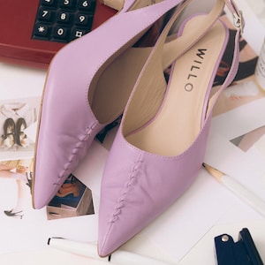 May include: A pair of lavender slingback kitten heels with a pointed toe and a decorative braided seam detail running down the center of the vamp. The shoes feature an adjustable ankle strap with a silver buckle and the brand name WILLO printed.