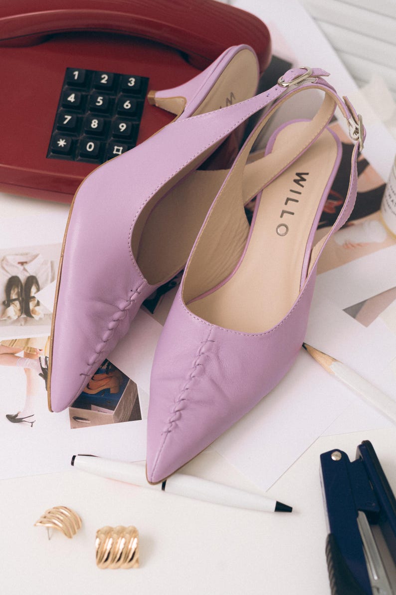 May include: A pair of lavender slingback kitten heels with a pointed toe and a decorative braided seam detail running down the center of the vamp. The shoes feature an adjustable ankle strap with a silver buckle and the brand name WILLO printed.