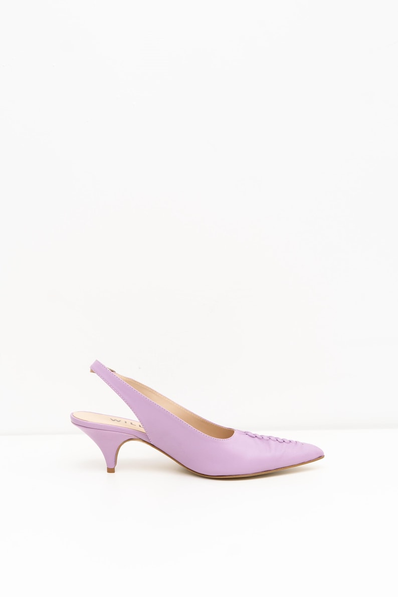 May include: A lavender slingback pump with a pointed toe and a low kitten heel. The shoe features a smooth finish, a delicate strap wrapping around the heel, and a subtle stitched detail along the top of the toe box for a clean, structured look.