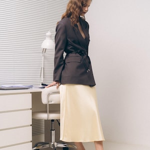 May include: A dark brown blazer with a black waist tie, a pale yellow midi skirt, and lavender pointed-toe slingback heels. The blazer features notched lapels and flap pockets, while the skirt has a smooth, satin-like finish and a straight hem.