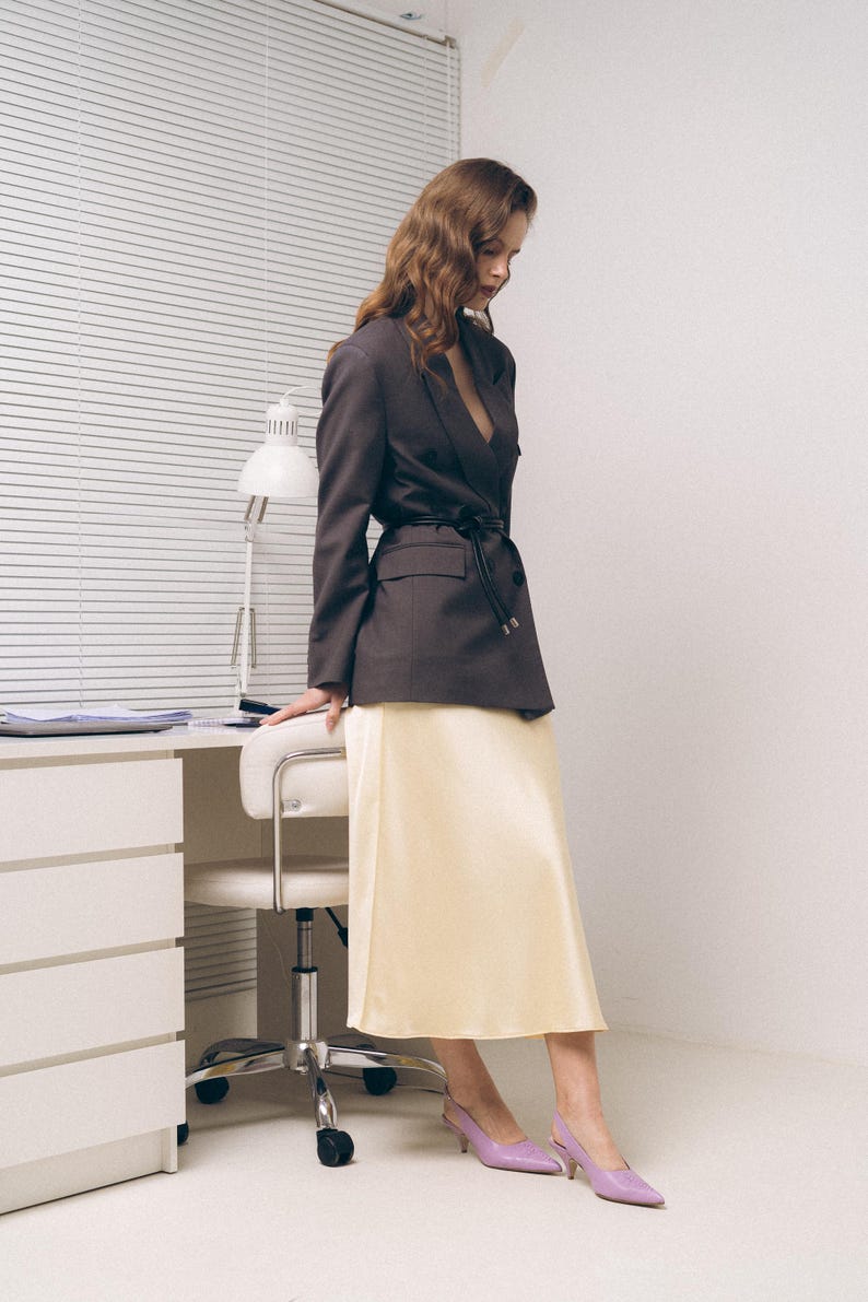 May include: A dark brown blazer with a black waist tie, a pale yellow midi skirt, and lavender pointed-toe slingback heels. The blazer features notched lapels and flap pockets, while the skirt has a smooth, satin-like finish and a straight hem.