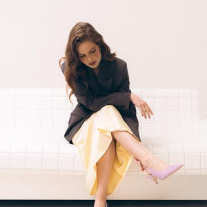 May include: A dark charcoal blazer paired with a pale yellow midi skirt and lavender pointed-toe slingback heels. The blazer features a structured collar and long sleeves, while the skirt has a smooth, flowing drape and a mid-calf length.