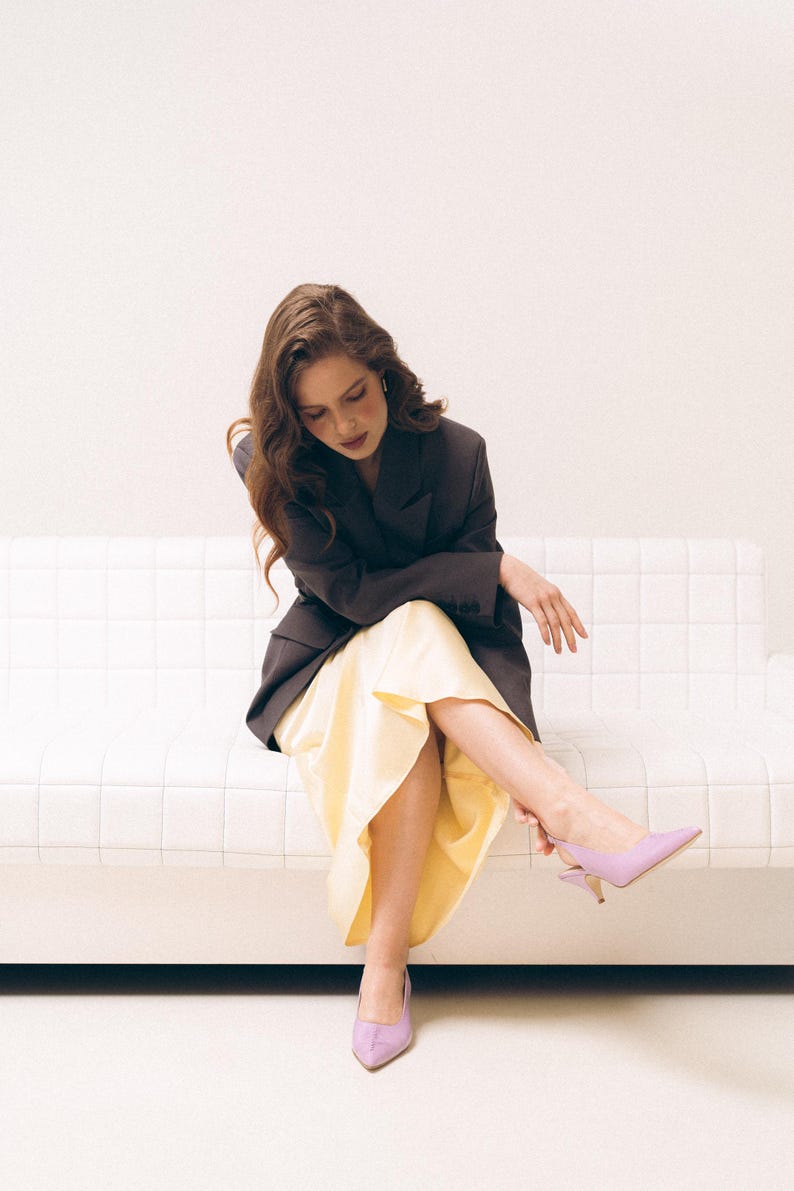 May include: A dark charcoal blazer paired with a pale yellow midi skirt and lavender pointed-toe slingback heels. The blazer features a structured collar and long sleeves, while the skirt has a smooth, flowing drape and a mid-calf length.