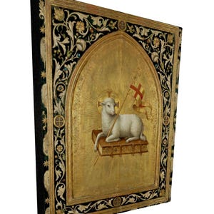 May include: A framed religious artwork featuring a lamb holding a flag with a cross. The lamb rests on a book within a gold arched frame. The frame has a black border with floral and leaf designs.