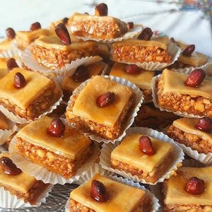 May include: A pile of golden-brown pastries, each square piece topped with a single nut. The pastries sit in white paper cups on a clear glass plate. The dessert has a layered appearance with a nutty filling.