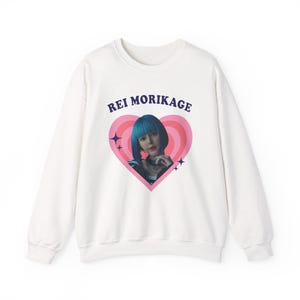 Könnte beinhalten: Weißes Sweatshirt mit einem Aufdruck einer Frau mit blauen Haaren in einem rosa Herzen, mit dem Text "REI MORIKAGE" darüber. Das Design beinhaltet Sterne. Ein lässiges Unisex-Kleidungsstück.
