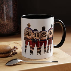 Peut inclure: Mug en céramique blanche avec intérieur et anse noirs. Le mug présente un motif de quatre joueurs de hockey en maillots vintage avec noms et numéros. Les joueurs portent des uniformes crème, rouges et bleus.