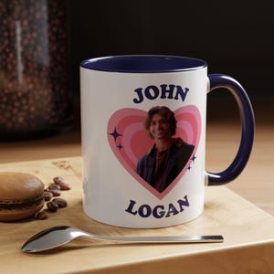 Może przedstawiać: Biały ceramiczny kubek z granatowym wnętrzem i uchwytem. Kubek przedstawia zdjęcie osoby w różowym sercu z napisem "JOHN" u góry i "LOGAN" na dole. Kubek stoi na drewnianej powierzchni z łyżką i makaronikiem.