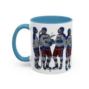 Puede incluir: Taza de cerámica blanca con interior y asa azules. La taza presenta un gráfico de cuatro jugadores de hockey con uniformes con la palabra "Briar" y números. Los jugadores llevan cascos y sujetan palos de hockey. La taza es de tamaño estándar.