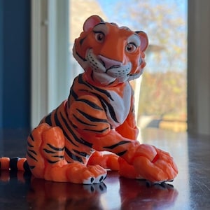 May include: Orange and black tiger figurine with white accents. The tiger is sitting with its paws crossed and has a friendly expression.