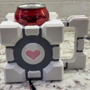 May include: White ceramic mug shaped like a cube with a gray heart and a handle. A red can is sitting inside the mug.