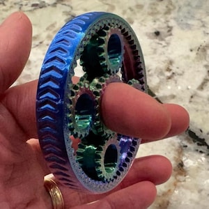 Puede incluir: Un mecanismo de engranajes multicolor impreso en 3D. El anillo exterior es azul con crestas en forma de cheurón. Los engranajes internos son verdes y turquesa. El mecanismo se sostiene en una mano.