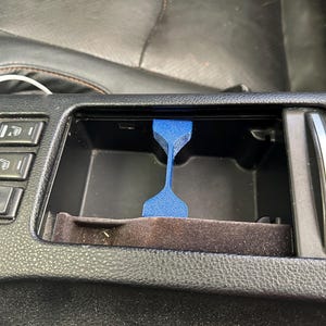 以下が含まれることがあります： 車のセンターコンソール用の青い3Dプリントの携帯電話ホルダー。ホルダーは、2つのカップホルダーの間に収まるように設計されており、携帯電話を垂直に保ちます。