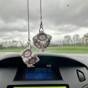 May include: Two white and pink cube-shaped car air fresheners hanging from the rearview mirror of a car. The cubes have a heart-shaped design on them.