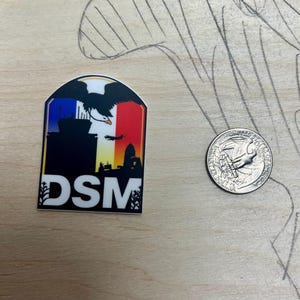 May include: A sticker with a black silhouette of a city skyline, an eagle flying over a building, and the text "DSM" in white letters. The sticker has a red, white, and blue background.