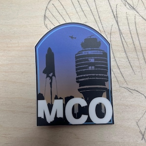 May include: A blue and white sticker with a silhouette of a space shuttle and a control tower. The sticker says "MCO" in white letters.