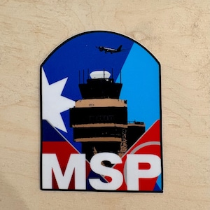 May include: A colorful patch featuring an airport control tower, a plane, and the letters "MSP" in white. The design incorporates blue, red, and white elements, with a star-like shape on the left side. The patch has a black border.