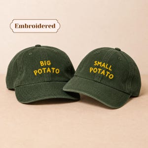 May include: Two dark green corduroy baseball caps featuring yellow embroidered text. One cap displays the words BIG POTATO and the other displays SMALL POTATO. The word Embroidered is printed in a decorative frame above the caps.