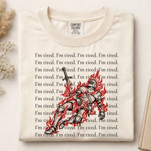 May include: A cream-colored t-shirt featuring a graphic of a knight in armor surrounded by red flames and holding a sword. The background text repeats the phrase I'm tired multiple times in black font. The neck label reads Comfort Colors.