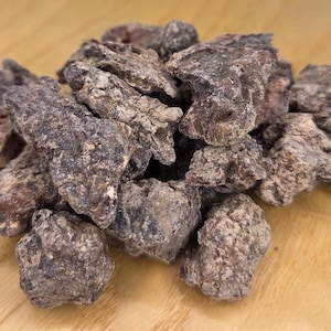 May include: A pile of dark brown, irregularly shaped pieces, possibly dried fruit or a similar food item. The pieces have a rough, textured surface and vary in size. The background is a light brown wooden surface.