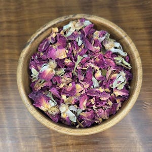 May include: A wooden bowl filled with dried rose petals. The petals are a mix of deep pink, purple, and faded yellow, with some green leaves. The bowl is made of light-colored wood and sits on a brown wooden surface.