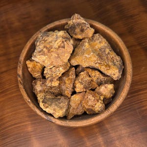 May include: A wooden bowl filled with irregularly shaped, amber-colored resin pieces. The resin has a rough, textured surface and varying shades of brown and gold. The bowl is made of light-colored wood and sits on a dark wooden surface.