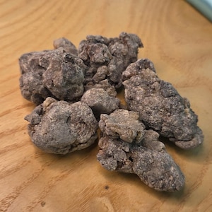 May include: A pile of irregularly shaped, dark brown, and textured pieces, possibly food items, are arranged on a light brown wooden surface. The items have a rough, granular appearance.