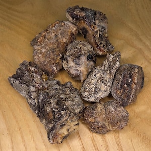 May include: A collection of irregularly shaped, dark brown pieces of frankincense resin, with lighter, dusty patches. The pieces vary in size and texture, resting on a light wood surface.