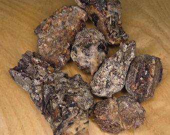 May include: A collection of irregularly shaped, dark brown pieces of frankincense resin, with lighter, dusty patches. The pieces vary in size and texture, resting on a light wood surface.