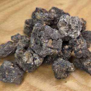 May include: A pile of dark, irregularly shaped rocks with a rough, textured surface. The rocks are primarily dark gray with lighter, tan-colored patches. They are arranged on a light brown wooden surface.