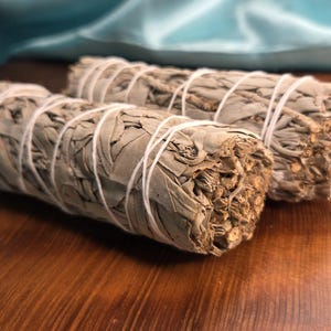 May include: Two bundles of dried white sage, tightly wrapped with white string. The sage has a light green color and a textured appearance. The bundles are arranged on a dark wooden surface, with a soft blue fabric in the background.