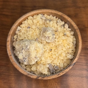 May include: A wooden bowl filled with light yellow, granular resin and several larger, irregularly shaped pieces. The resin has a slightly translucent appearance, with some darker flecks. The bowl sits on a dark wooden surface.