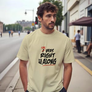 May include: A cream-coloured t-shirt featuring the phrase "SHE WAS RIGHT ALL ALONG" in black and red lettering. The t-shirt has a crew neck and is made of a soft fabric. The text is in a bold, handwritten style.
