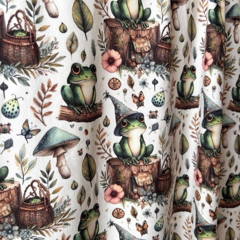 A close-up of draped fabric shows a repeating pattern of green frogs in wizard hats on tree stumps, surrounded by mushrooms and leaves.